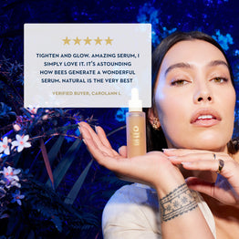 A woman holds a small amber serum bottle in her hand against a dark blue floral background, with a large review card overlaid on the left showing a five-star quote and the text 'Verified Buyer, Carolynn L.'.